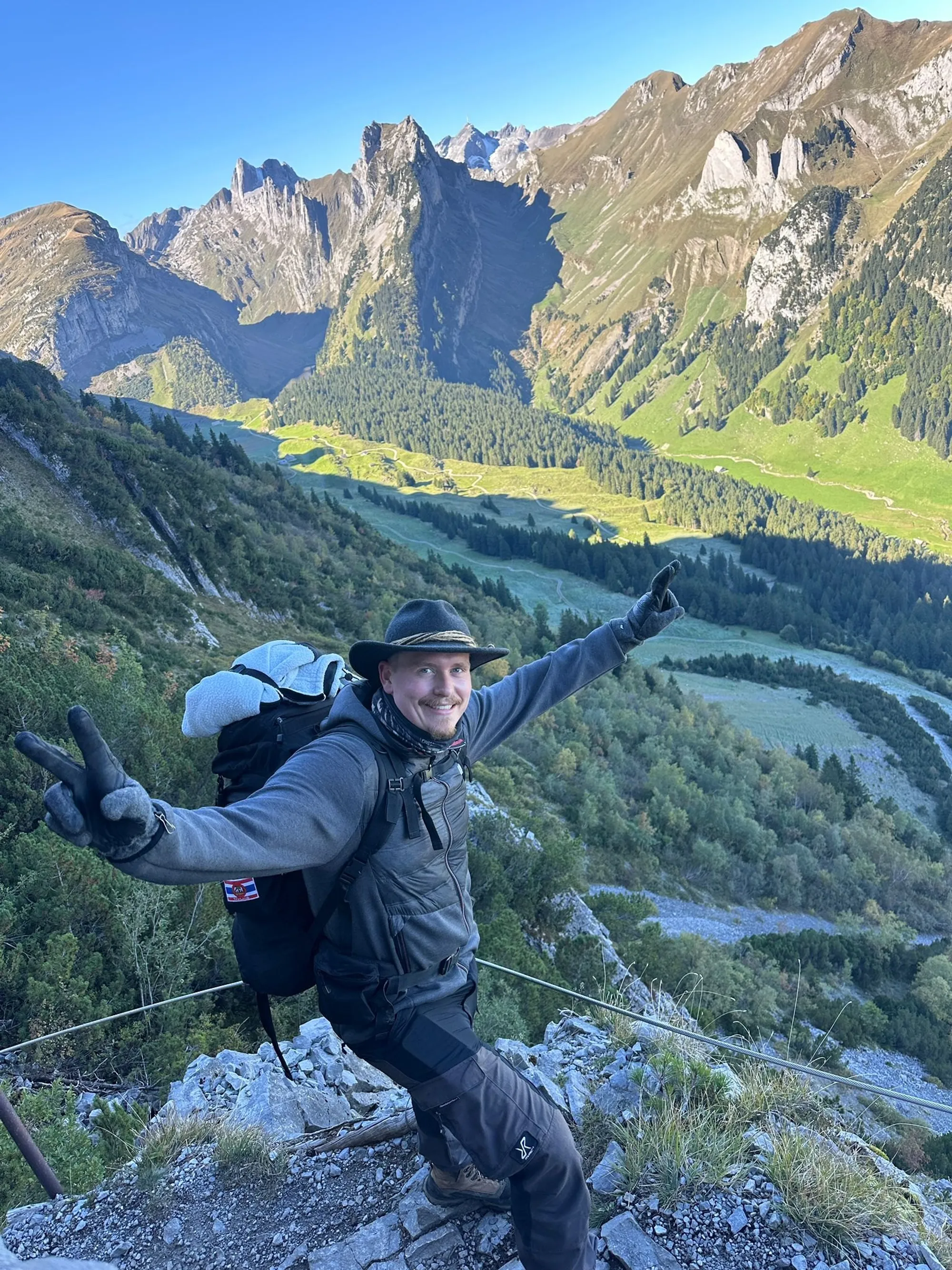 Max auf einem Bergpfad, Arme ausgebreitet, im Hintergrund ein grünes Alpental